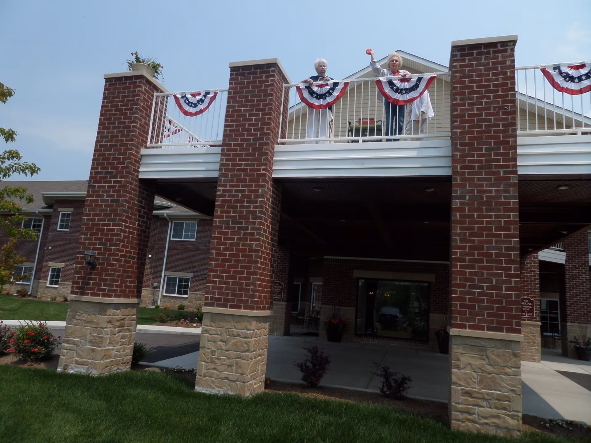 Front entrance of a brick building with large columns and a balcony decorated with red-white-blue bunting where two people are standing and waving.