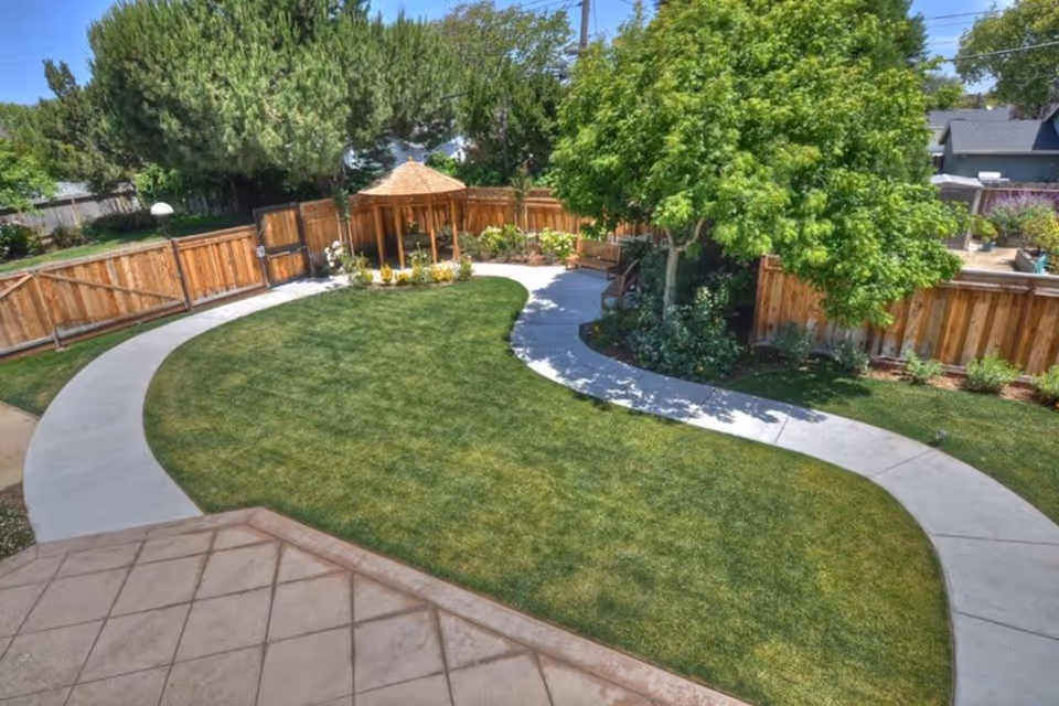 A well-maintained outdoor garden area with a curved concrete pathway surrounding a green lawn. There is a wooden gazebo with a shingled roof in the background, surrounded by a wooden fence and various trees and shrubs providing shade and greenery.