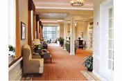 Well-lit carpeted common hallway in a senior living facility with seating areas, columns, plants, and large windows.
