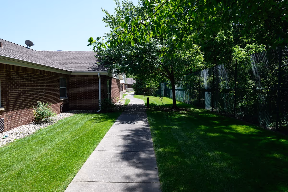 A sunny outdoor walkway beside a brick building with well-maintained green grass and trees. A black chain-link fence runs along the right side of the path, with shadows cast on the grass and sidewalk.