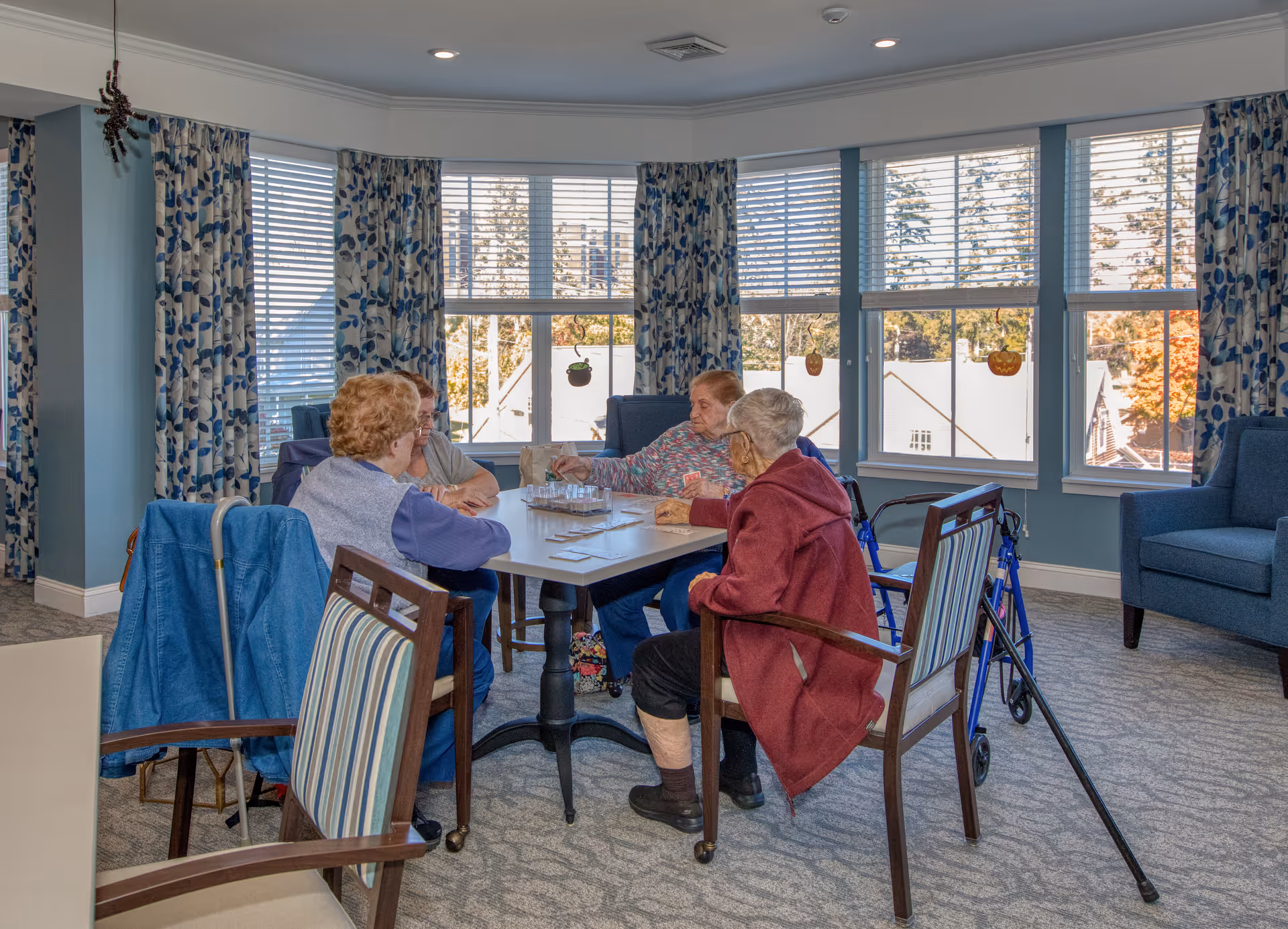 Four elderly women sitting around a square table in a bright room with large windows and blue patterned curtains. The room has light blue walls, carpeted floor, and comfortable chairs. Two walkers are visible near the women.