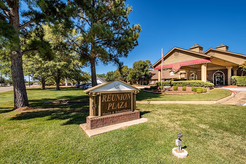 Front entrance of Reunion Plaza Senior Care Center with a prominent sign on a grassy lawn and the facility building in the background under a blue sky.