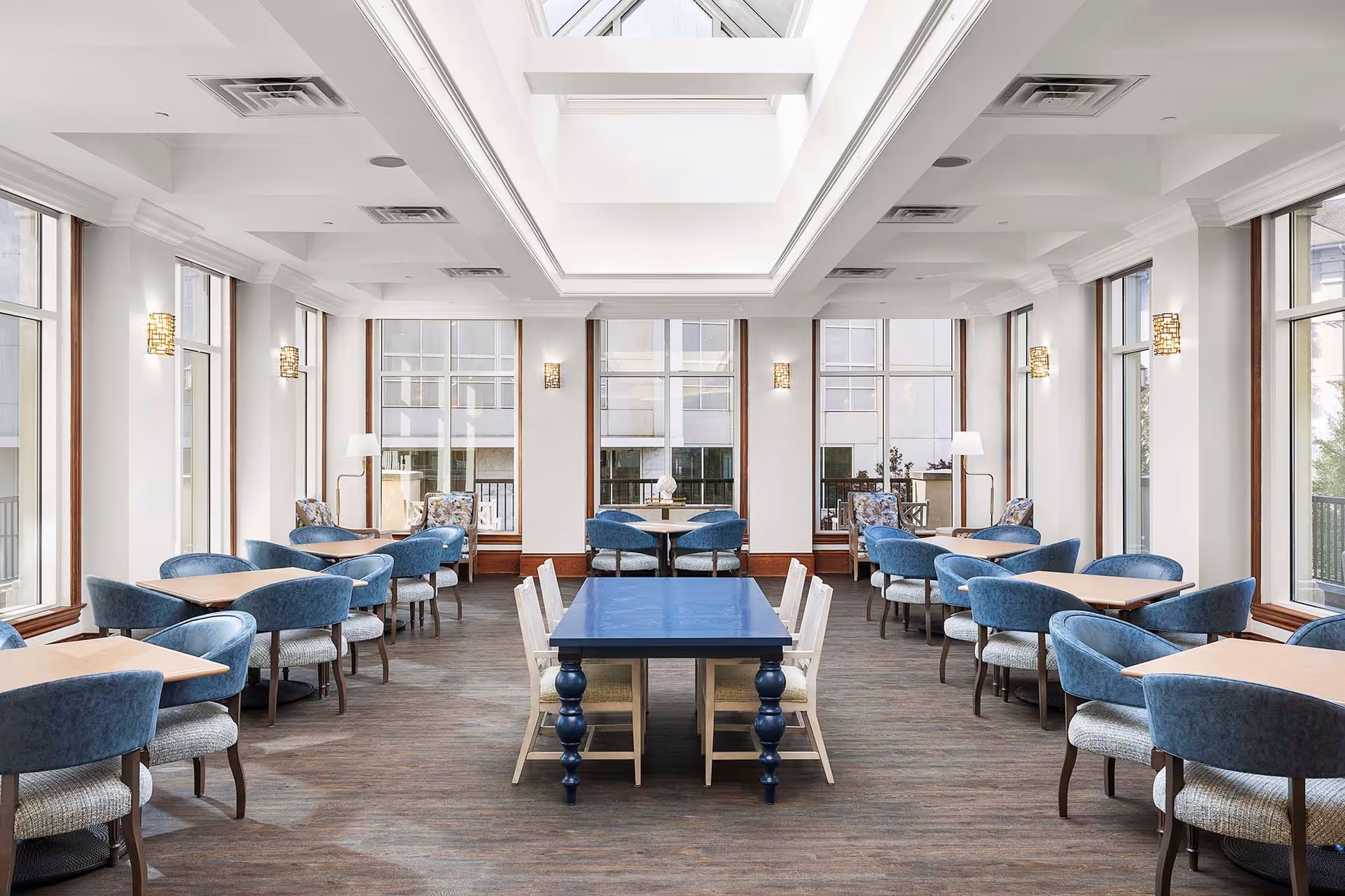 Bright dining room with multiple tables and blue upholstered chairs arranged neatly. The room features large windows on three sides allowing natural light to fill the space, and a skylight in the ceiling. The walls are white with decorative sconces, and the floor is covered with a patterned carpet.