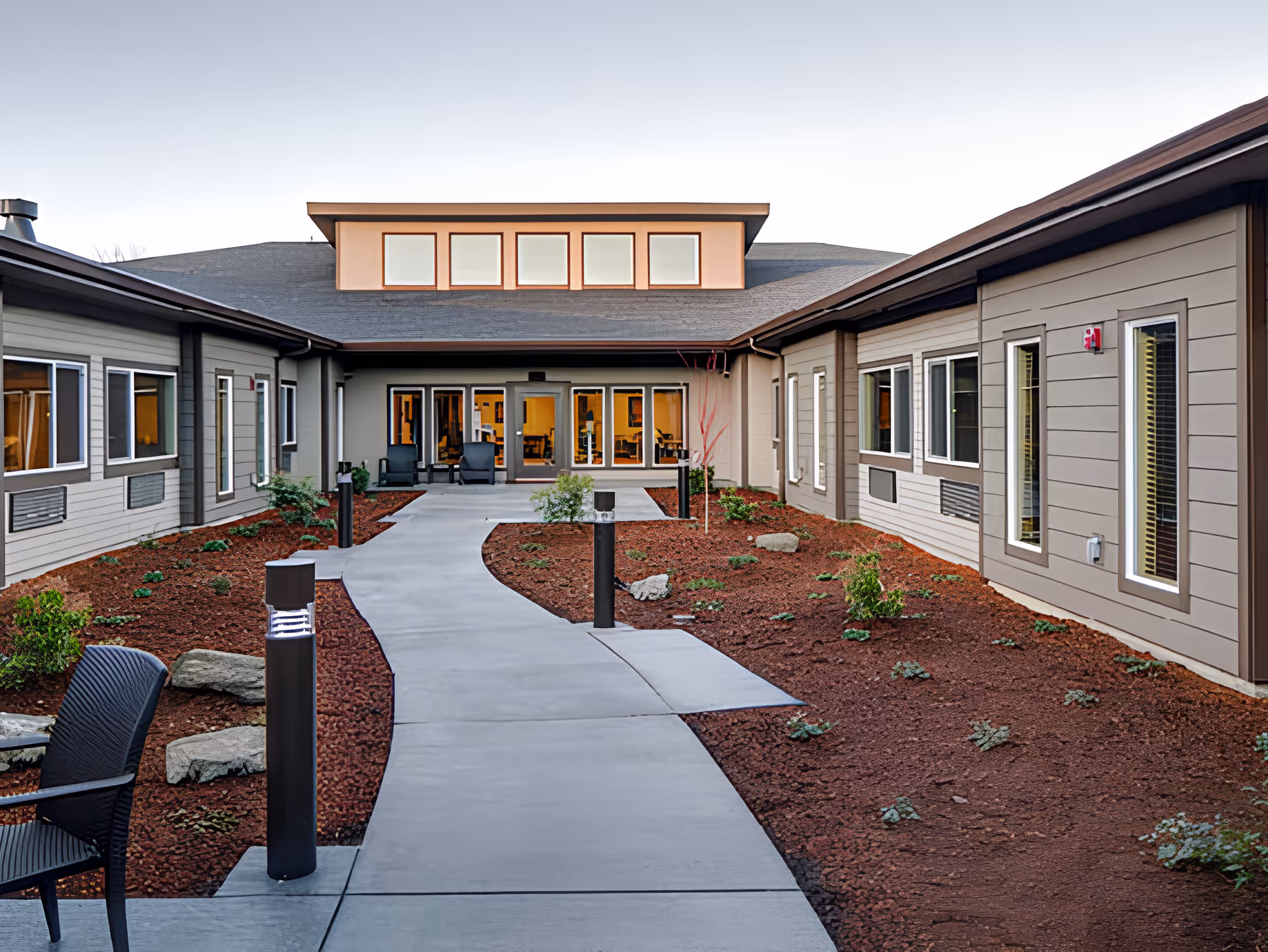 Outdoor courtyard area of Avamere Transitional Care of Puget Sound with a curved concrete walkway, small plants and shrubs in mulched beds on either side, modern building exterior with multiple windows, and patio chairs near the entrance.