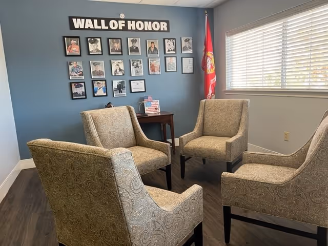 Four upholstered chairs arranged in a small seating area facing a 'WALL OF HONOR' photo display on a blue wall next to a window with blinds.