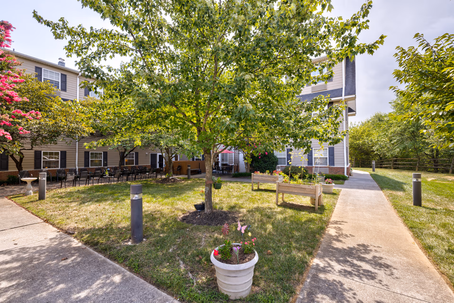 Outdoor courtyard area at TerraBella Pheasant Ridge featuring a large tree in the center, flower pots, raised garden beds, and a paved walkway. The courtyard is surrounded by a two-story building with windows and black shutters, and there are several chairs lined up along the building's wall. The area is well-lit with outdoor lamps and has green grass and other trees providing shade.
