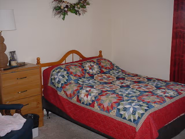 A bedroom with a bed covered by a colorful patchwork quilt, wooden headboard, a dresser with a lamp, and red curtains.