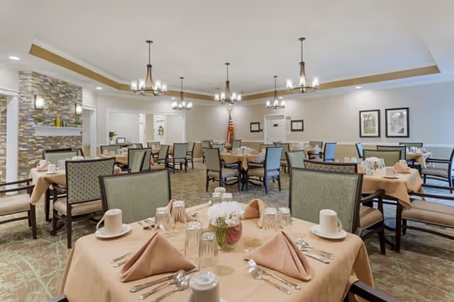 A spacious dining room with multiple tables covered in beige tablecloths, each set with folded napkins, cups, glasses, and silverware. The room features comfortable chairs with patterned upholstery, a stone fireplace on the left, and framed artwork on the walls. Several chandeliers hang from the ceiling, providing warm lighting.