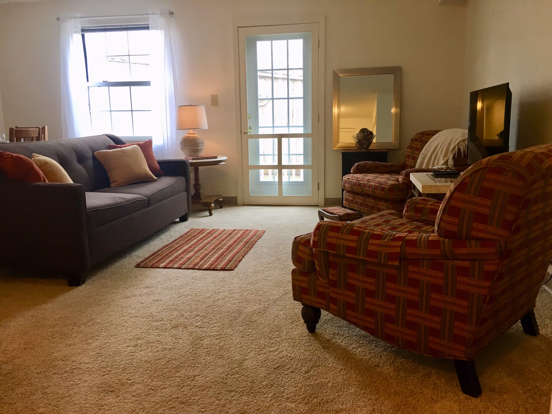 A cozy living room with a gray sofa adorned with beige and red pillows on the left, a small round wooden side table with a lamp next to it, a striped red and beige rug on the carpeted floor, two patterned armchairs on the right, a small table with a TV, and a large mirror on a stand against the wall. A window with sheer white curtains and a glass door letting in natural light are visible in the background.