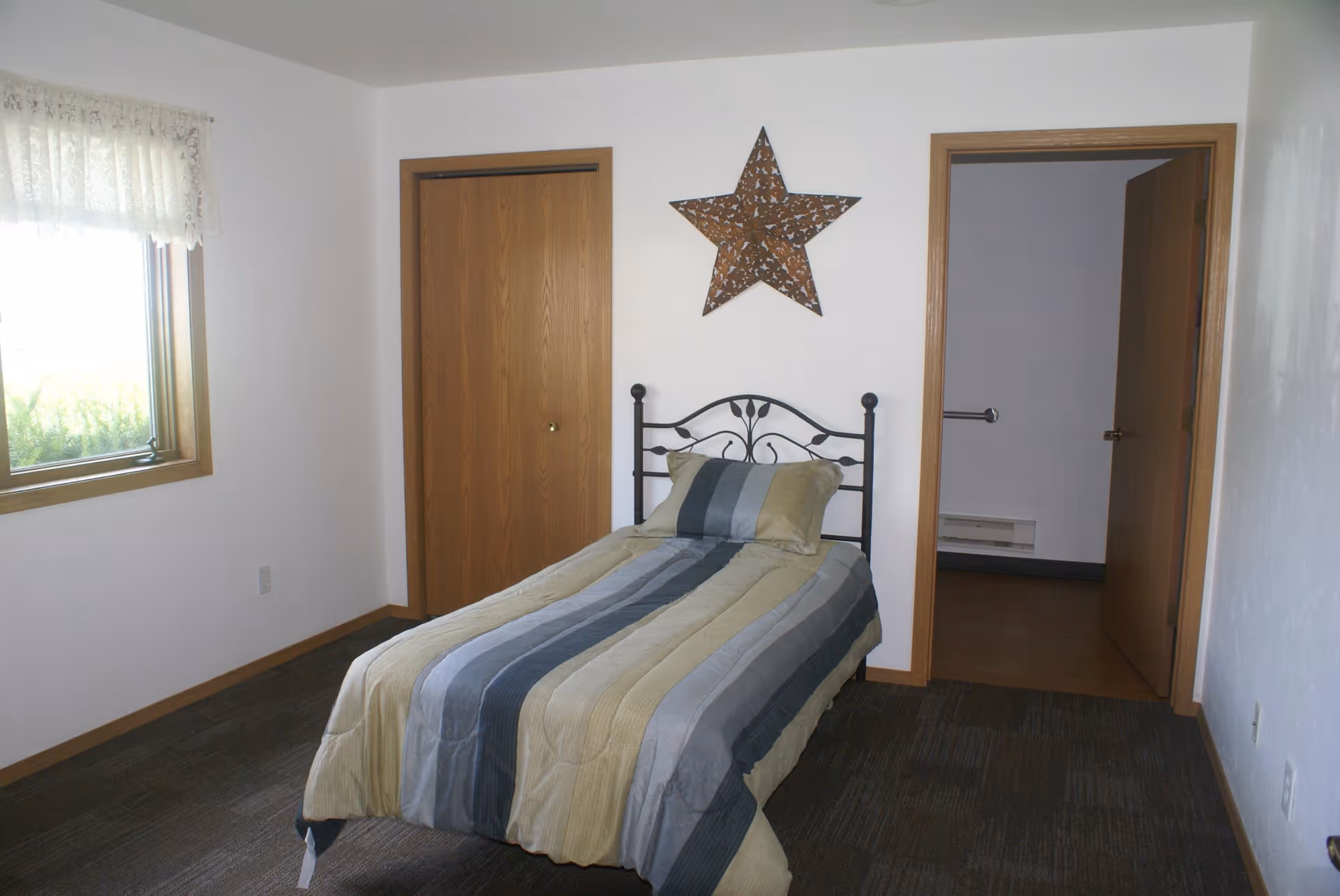A simple bedroom with a single bed covered in a striped comforter in shades of gray, blue, and beige. The bed has a black metal headboard and a matching pillow. On the wall above the bed is a large decorative star. To the left of the bed is a window with lace curtains, and to the right are two wooden doors, one slightly open showing a glimpse of another room.