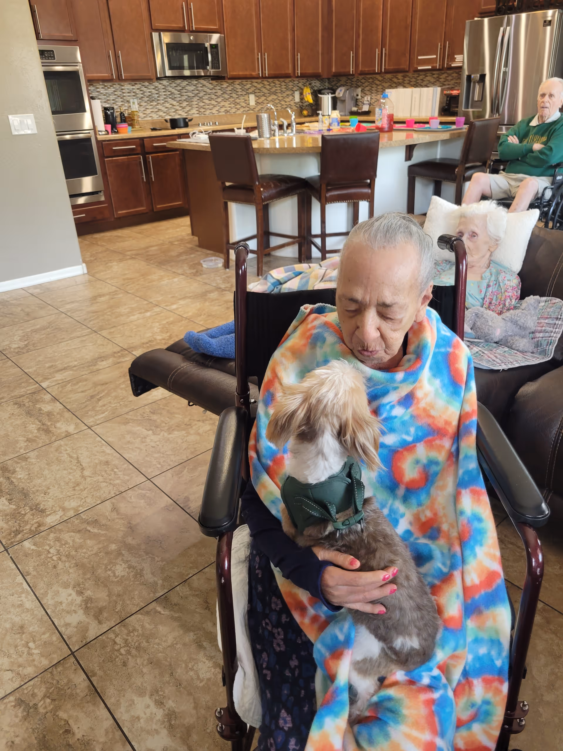 An elderly person in a wheelchair wrapped in a colorful blanket holds a small dog in a communal living area with a kitchen visible in the background.