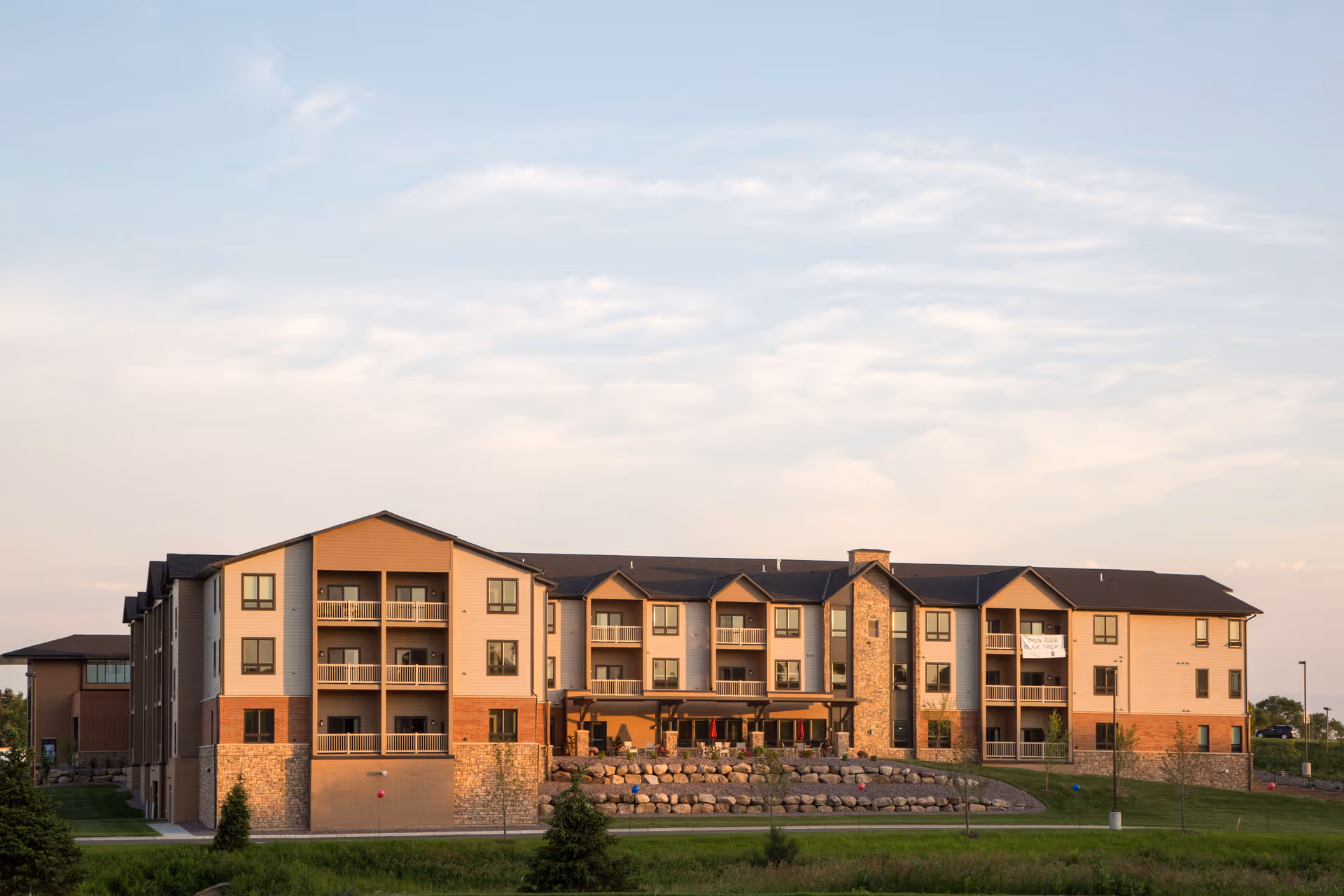 Exterior view of a multi-story senior living facility building with balconies, stone and brick accents, and a well-maintained lawn with small trees and rocks in front under a partly cloudy sky.