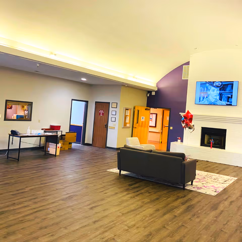 A spacious interior room with wood flooring, a dark gray couch and a patterned armchair facing a white brick fireplace with a mounted flat-screen TV above it. There are red and silver balloons near the fireplace. The walls are painted in neutral tones with one purple accent wall. A table with some boxes and supplies is against one wall, and several doors lead to other rooms.