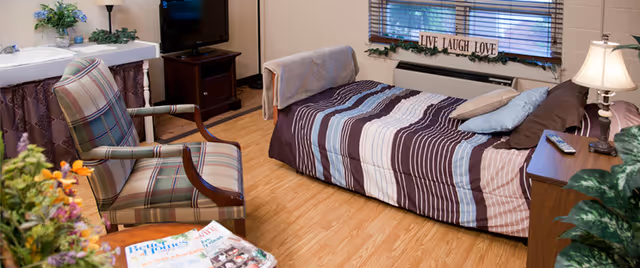 A cozy room with a single bed covered in a striped brown, blue, and white comforter, a wooden nightstand with a lamp and remote control, a plaid upholstered armchair, a small TV on a stand, a sink with a mirror, and a window with blinds and a decorative 'LIVE LAUGH LOVE' sign on the windowsill.