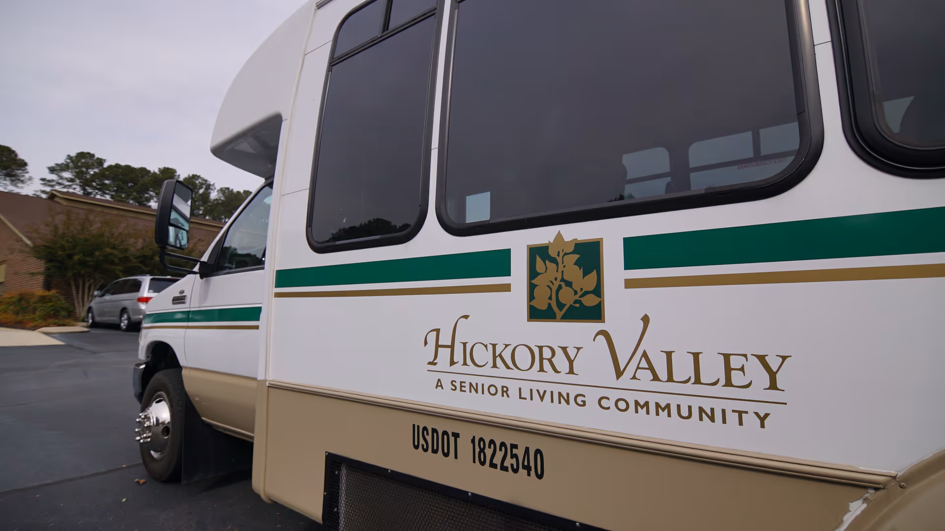 Side of a Hickory Valley senior living community shuttle bus with logo parked outside near a brick building.