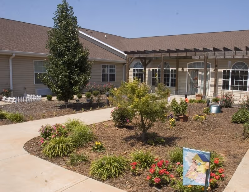 Outdoor garden area with a variety of plants and flowers, a concrete walkway, and a beige building with large windows and a pergola in the background under a clear sky.
