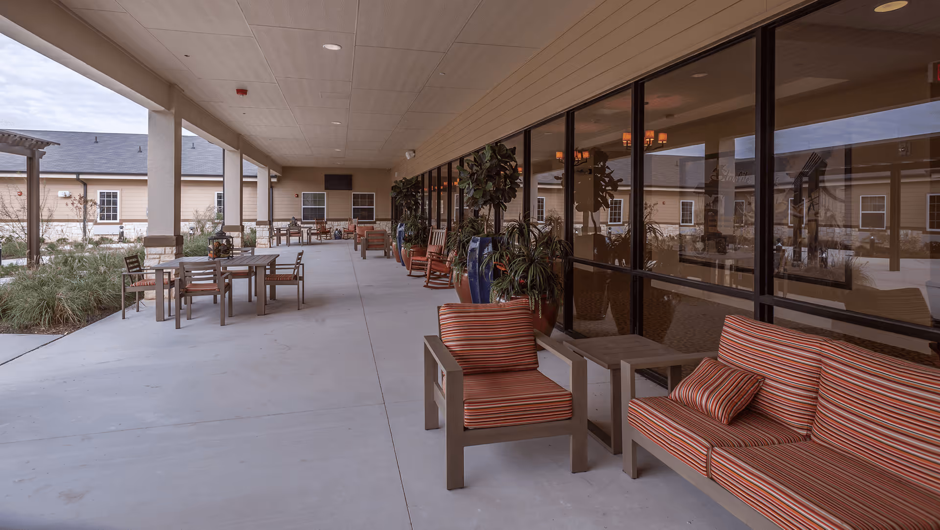 Covered outdoor patio area with multiple seating arrangements including striped cushioned chairs and sofas, tables, and potted plants along the glass windows of the building. The patio overlooks a garden area with grass and shrubs, and the building exterior is visible in the background.