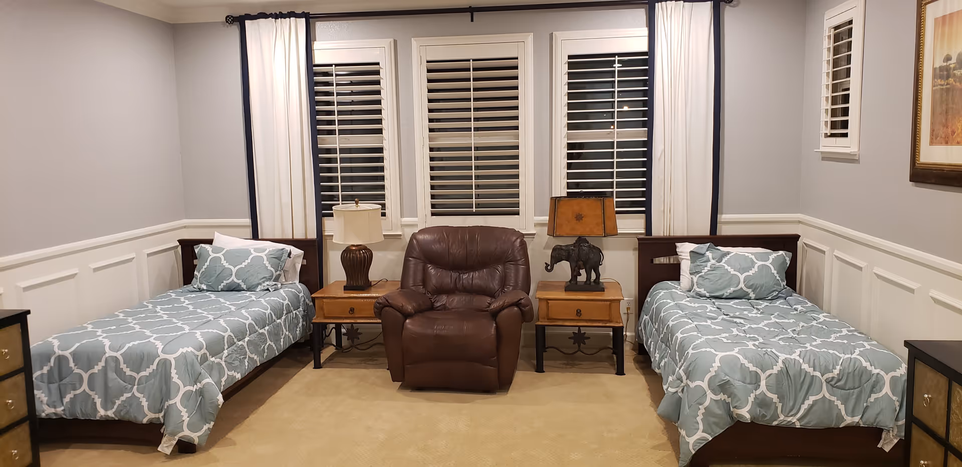 A bedroom with two twin beds on either side of a brown leather armchair. Each bed has a blue and white patterned comforter and pillows. Between the beds and armchair are two wooden nightstands with lamps, one lamp has a white shade and the other has a brown shade with an elephant base. Behind the armchair are three windows with white shutters and white curtains with dark trim. The walls are light gray with white wainscoting and there is a framed picture on the right wall.