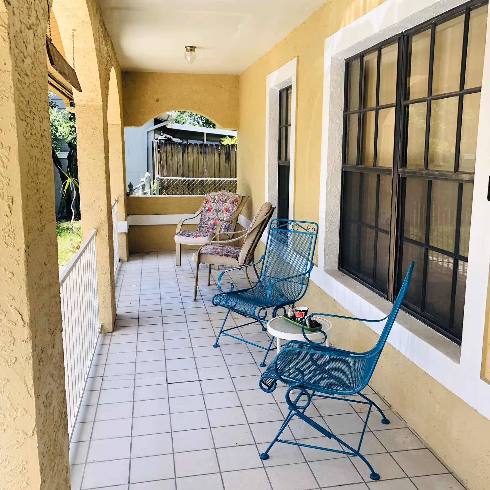 A covered outdoor porch area with yellow textured walls and white tiled floor. There are two blue metal chairs and a small white round table with a cup and small plants on it. Further back, there are two cushioned armchairs with floral patterned cushions. The porch has arched openings and a view of a wooden fence and greenery outside.
