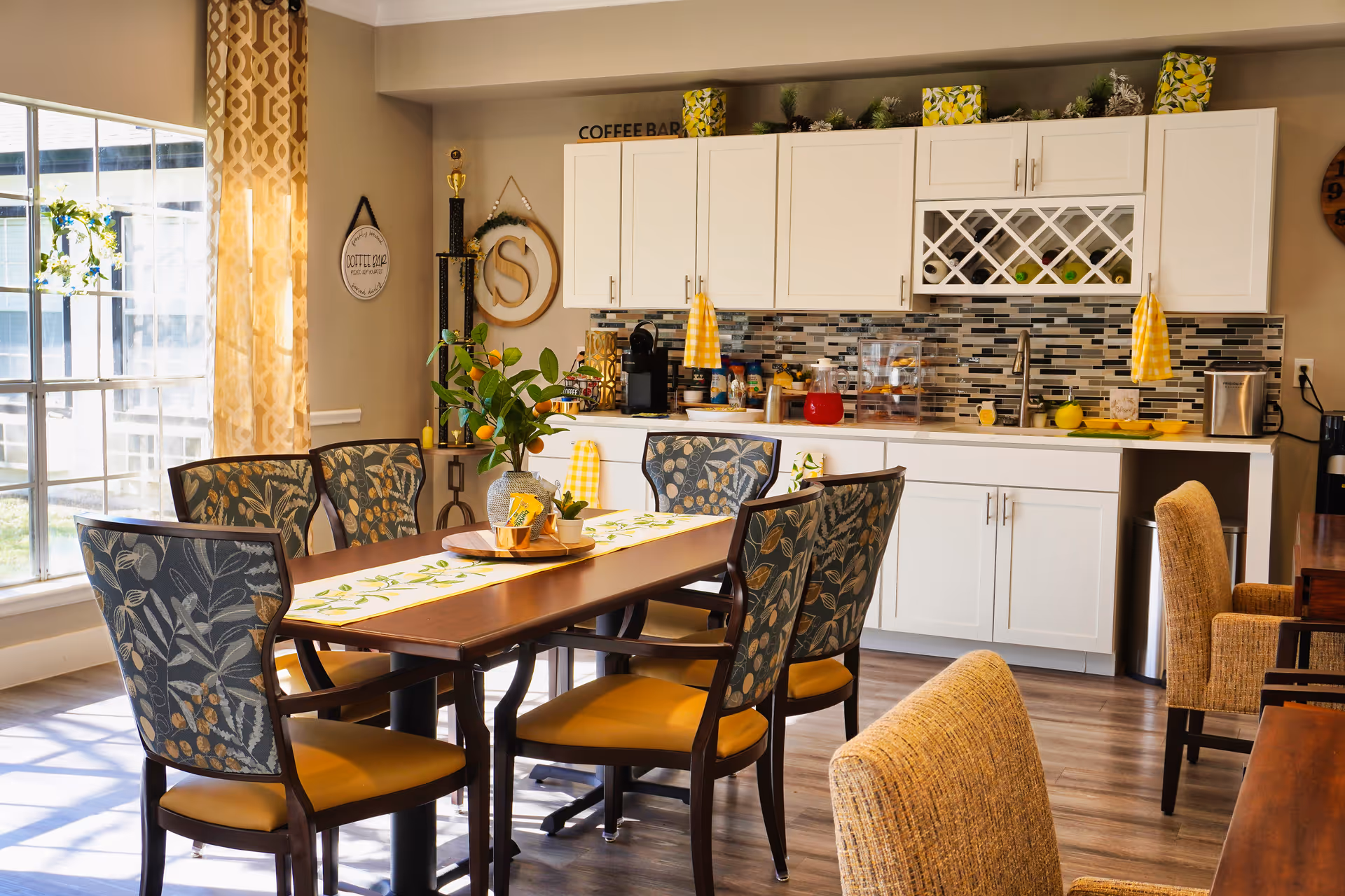 Bright communal dining area with a wooden table and patterned chairs in front of a white kitchenette and large window.