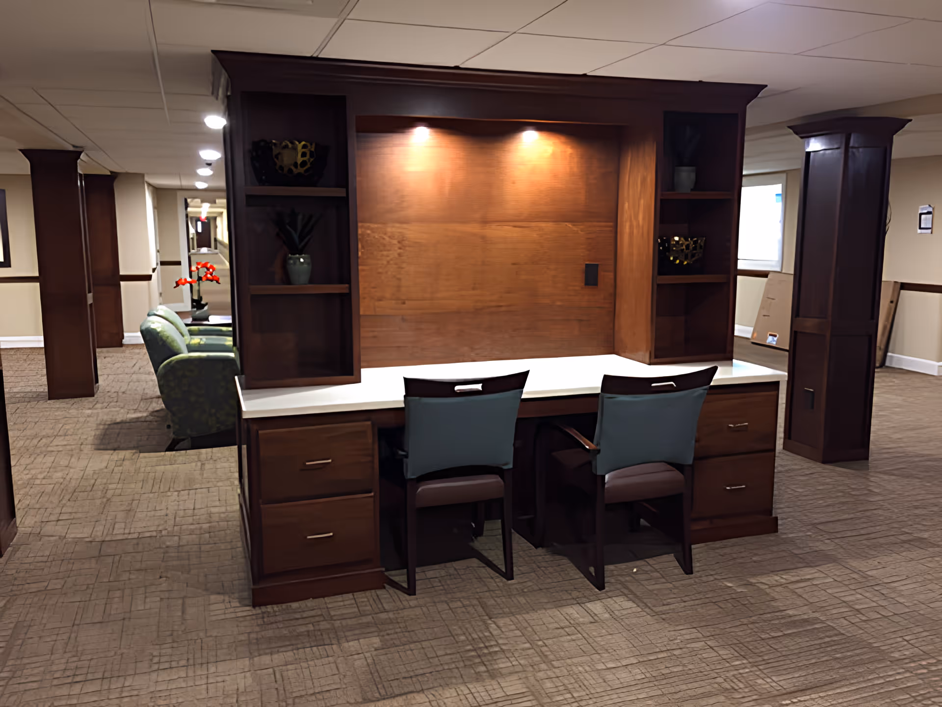 Interior view of a senior living facility hallway featuring a wooden desk with two chairs, built-in shelves with decorative items, and a seating area with green armchairs and a red flower arrangement in the background.