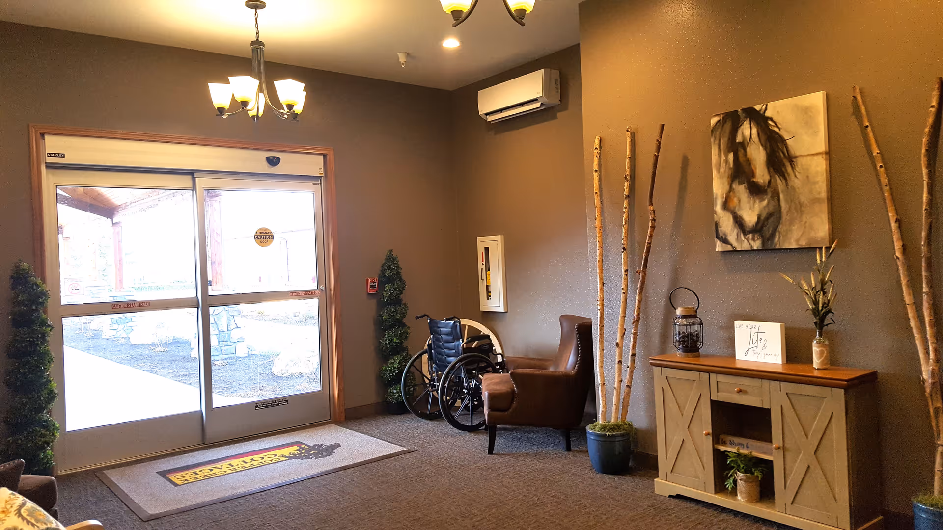 Interior view of an entrance area with automatic sliding glass doors, a brown armchair, a wheelchair, decorative plants, a wooden cabinet with decor items including a lantern, a small sign, and a vase with flowers, and a painting of a horse on the wall.