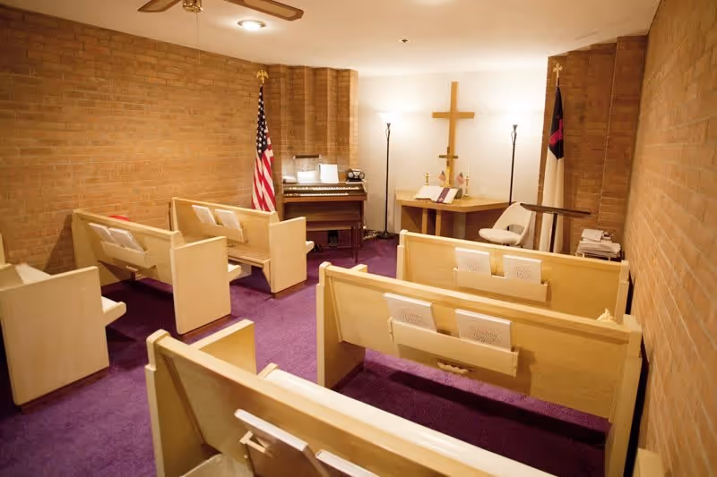 Small chapel room with wooden pews arranged facing a wooden altar with a cross on the wall behind it. There are two flags, an American flag and a Christian flag, placed on either side of the altar. A small organ is positioned against the back wall, and the floor is covered with purple carpet. The walls are made of light brown brick.