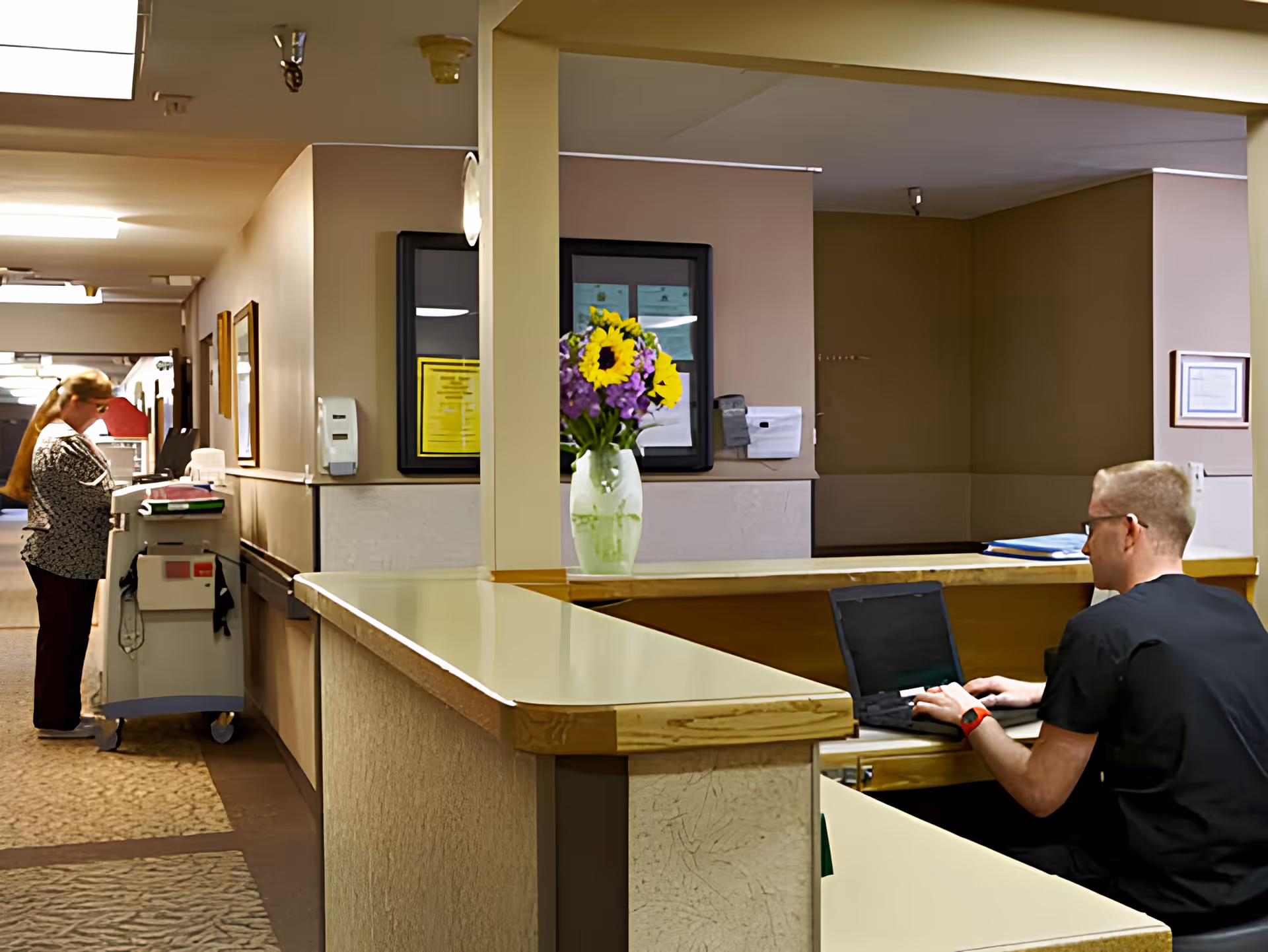 A reception/nurse station interior with a man using a laptop at the front desk, a vase of flowers on the counter, and a woman with a cart in the hallway.