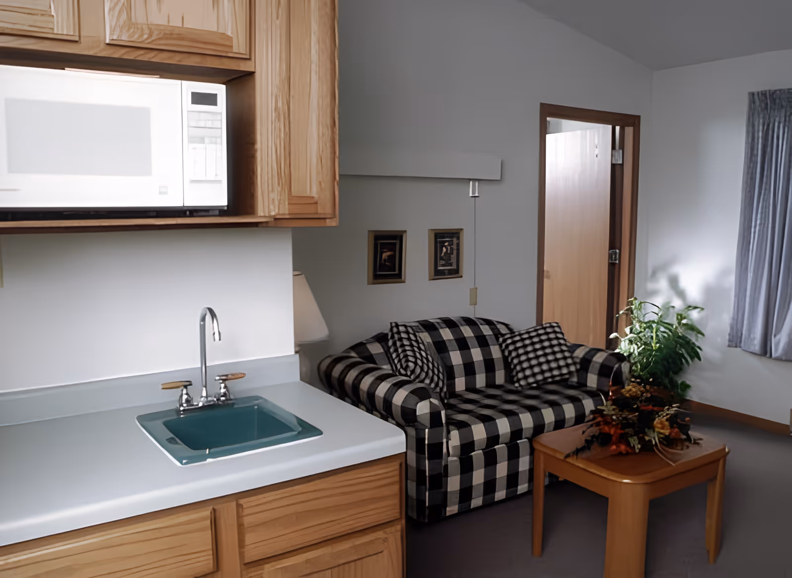 Small furnished living area with a black-and-white plaid loveseat, coffee table, and a kitchenette sink under wooden cabinets.