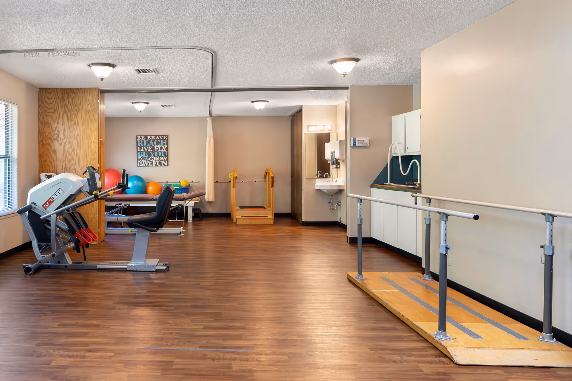 A rehabilitation room with exercise equipment including a recumbent bike, therapy balls, a treatment table, parallel bars for walking exercises, a small wooden step platform, a sink with a mirror, and cabinets. The room has wood flooring and beige walls with ceiling lights.