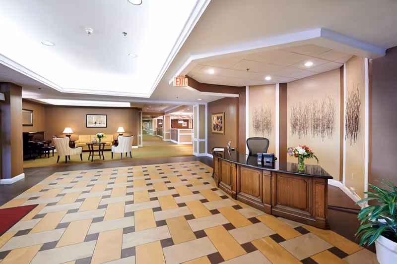 Reception area of a senior living facility with a wooden front desk on the right, decorated with a vase of flowers. The floor has a patterned tile design. In the background, there is a seating area with a round table, chairs, and lamps, along with a piano against the wall. The walls are painted in warm tones with framed artwork and decorative tree designs. Ceiling lights illuminate the space.
