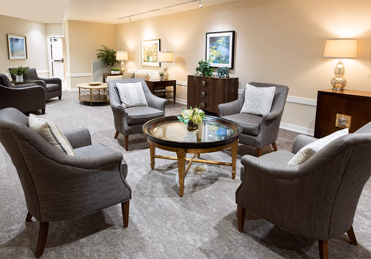 A cozy sitting area in a senior living facility with four gray upholstered armchairs arranged around a round glass-top coffee table. The room has beige walls, carpeted floor, decorative lamps on wooden side tables, framed artwork on the walls, and some green plants for decoration.