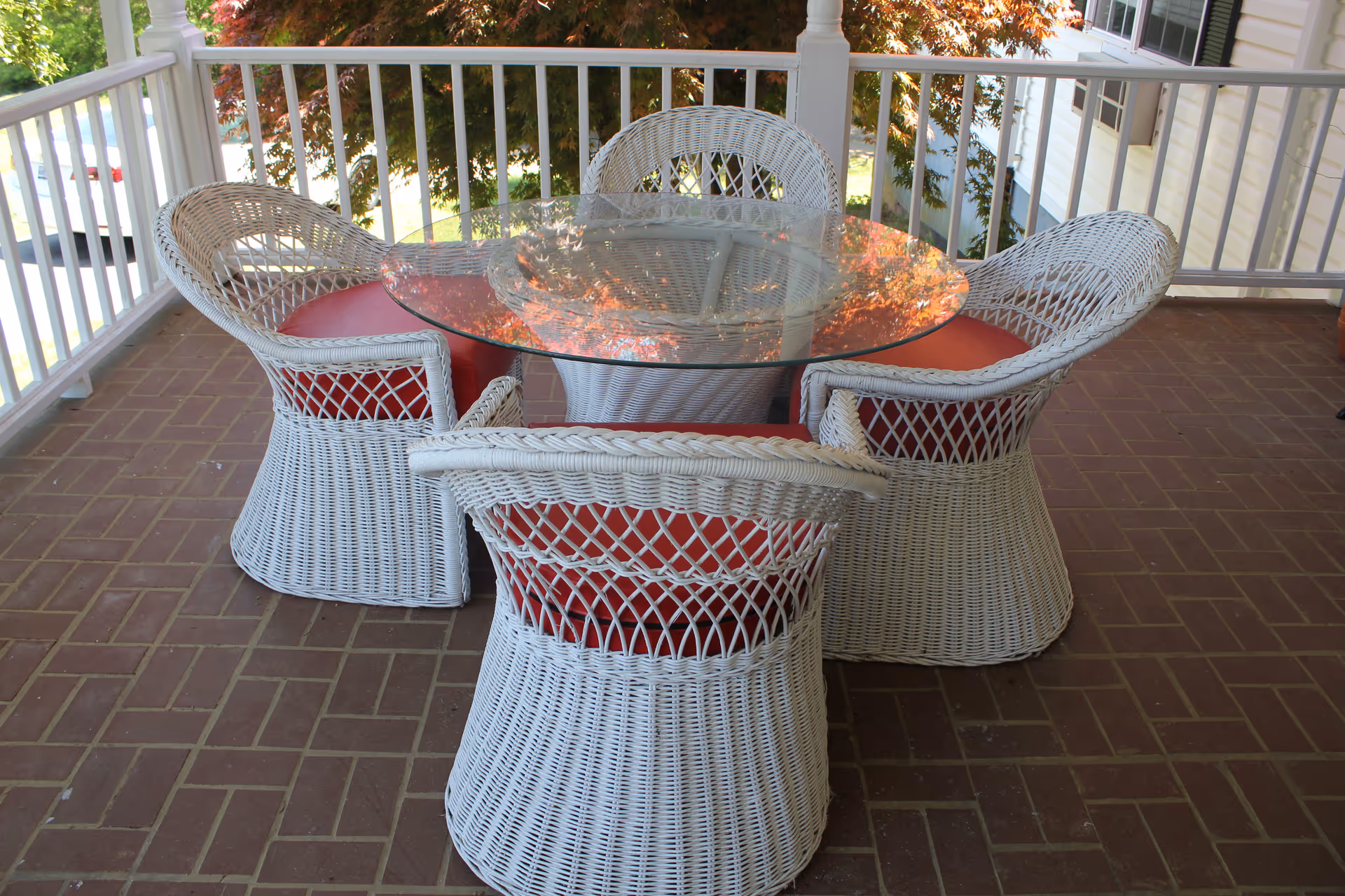 A covered outdoor patio area with a round glass-top table surrounded by four white wicker chairs with red cushions, set on a brick floor with white railing and trees visible in the background.