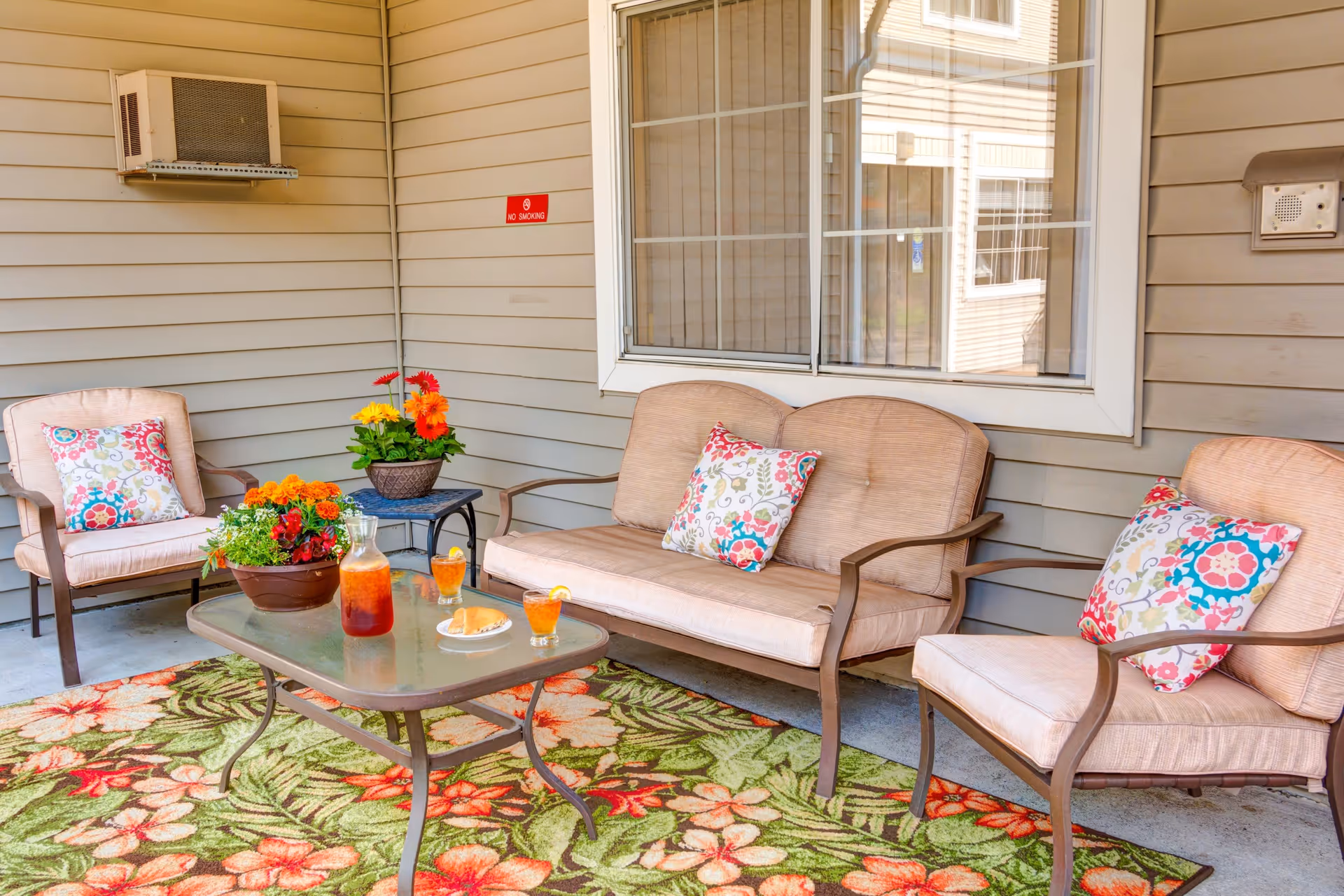 Outdoor patio area with cushioned seating including a loveseat and two chairs, each with colorful floral pillows. A glass-top table holds a pitcher of iced tea, two glasses with lemon slices, and a plate with a sandwich. There are potted flowers on a small side table and on the ground, and a floral patterned outdoor rug covers the floor. The area is adjacent to a building with beige siding and a window.