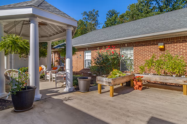 Outdoor patio area with a white gazebo featuring columns, wicker chairs with orange cushions, potted plants, raised garden beds with flowers and greenery, and a brick building with windows in the background under a clear blue sky.