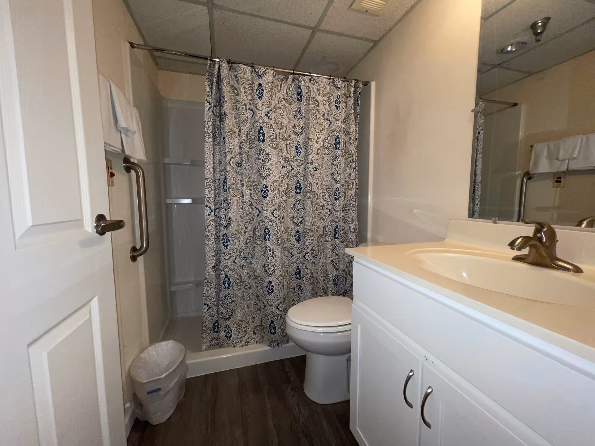 A bathroom with a white door, a shower with a patterned blue and white curtain, a white toilet, a white vanity with a sink and bronze faucet, a large mirror above the sink, a towel rack with white towels, and a small trash can on the floor.