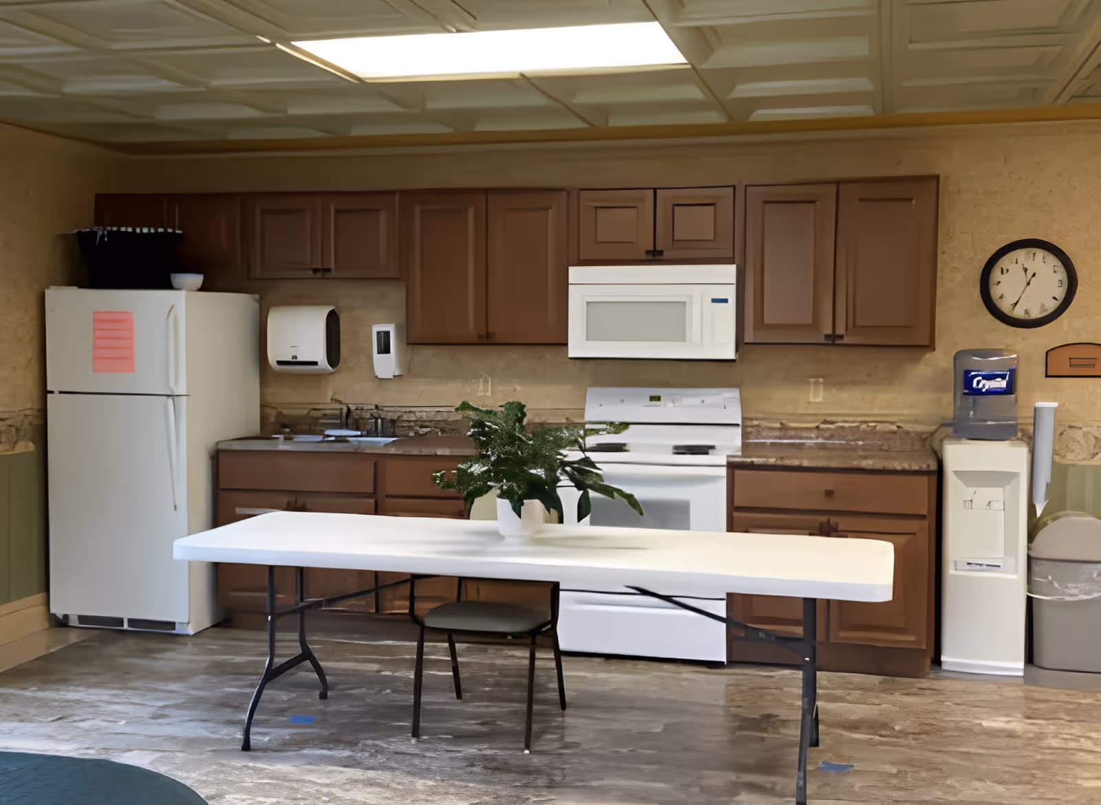 Interior view of a kitchen area with brown cabinets, a white refrigerator, a white microwave above a white stove, a water cooler, and a white folding table with a green plant in a white pot on top. A clock is mounted on the wall above the water cooler.