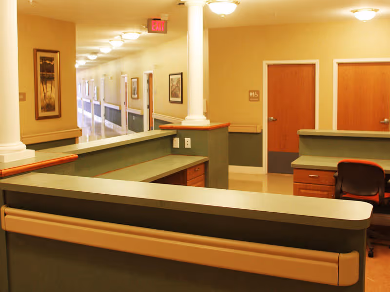 Reception/nurses' station with desks and chairs facing a long corridor lined with doors and framed artwork.