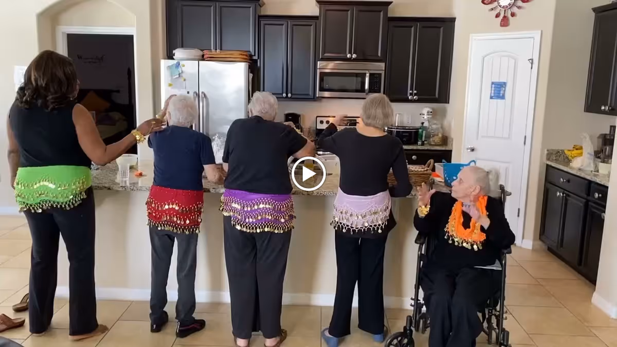 Five older adults and a caregiver wearing colorful hip scarves gathered at a kitchen counter, with one resident seated in a wheelchair.