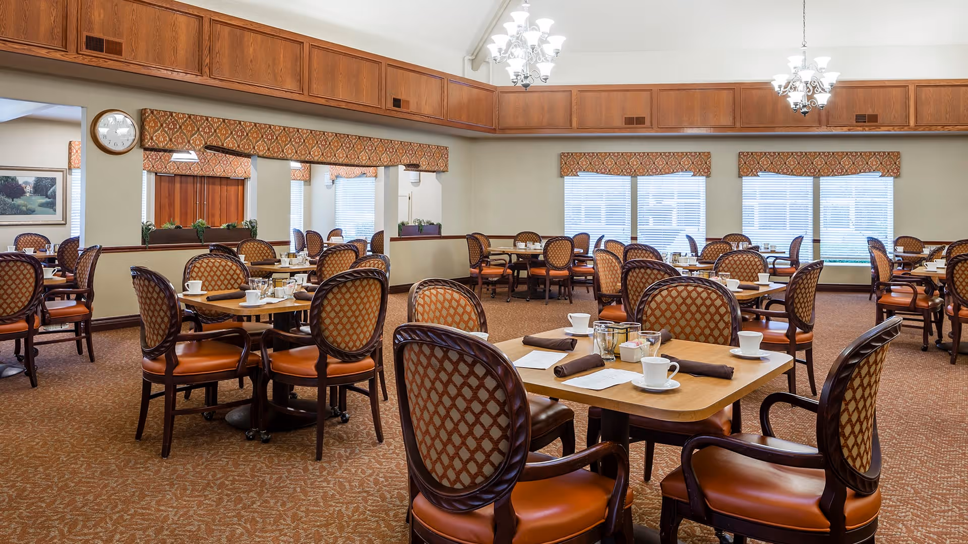 A spacious dining room with multiple wooden tables and cushioned chairs arranged neatly. Each table is set with white cups, saucers, napkins, and condiments. The room features large windows with patterned valances, chandeliers hanging from a high ceiling, and a carpeted floor with a warm, patterned design.
