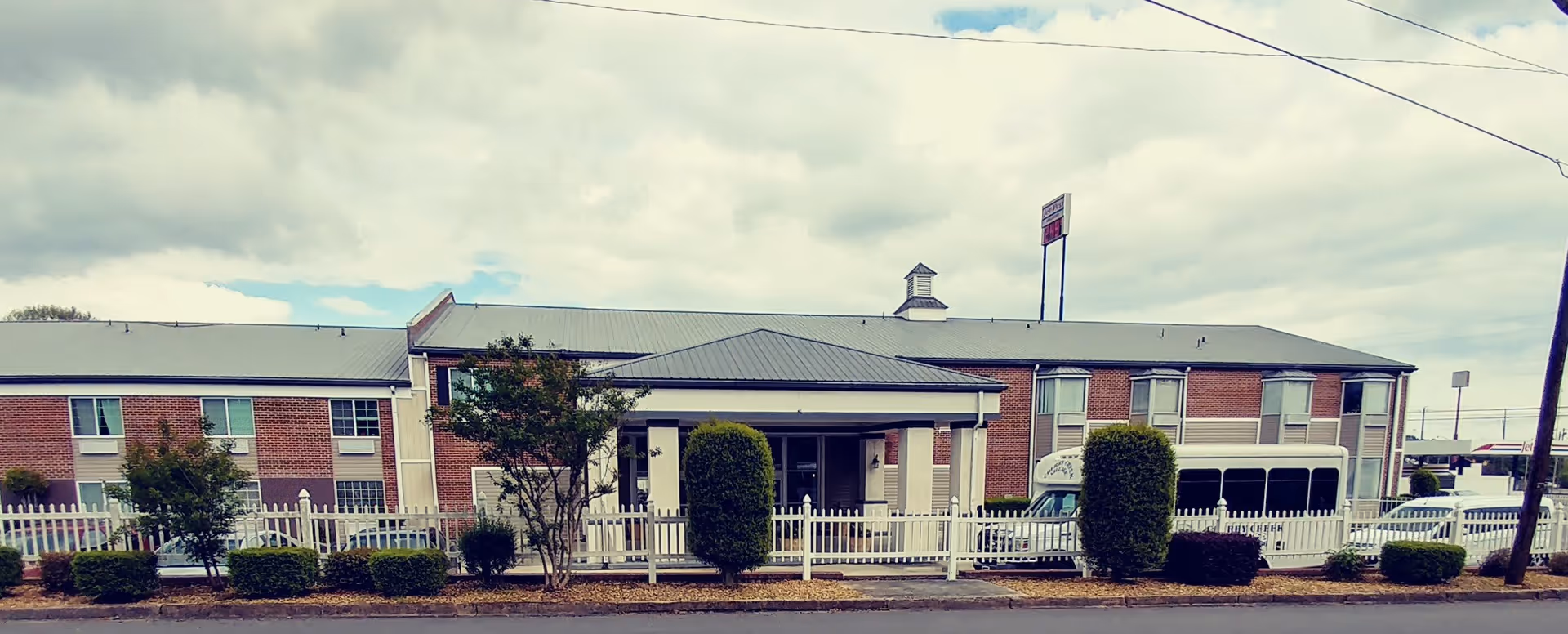 Exterior view of a two-story brick building with a gray roof, white picket fence, and neatly trimmed bushes in front under a cloudy sky.