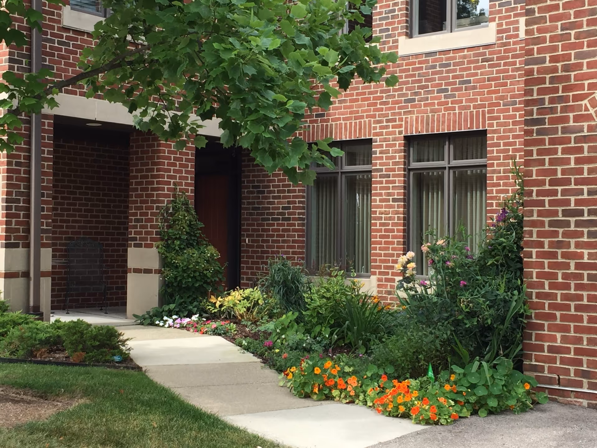 A brick building exterior with a concrete walkway leading to a covered entrance. There are green shrubs and colorful flowers planted along the walkway, and a tree with green leaves partially shading the area.