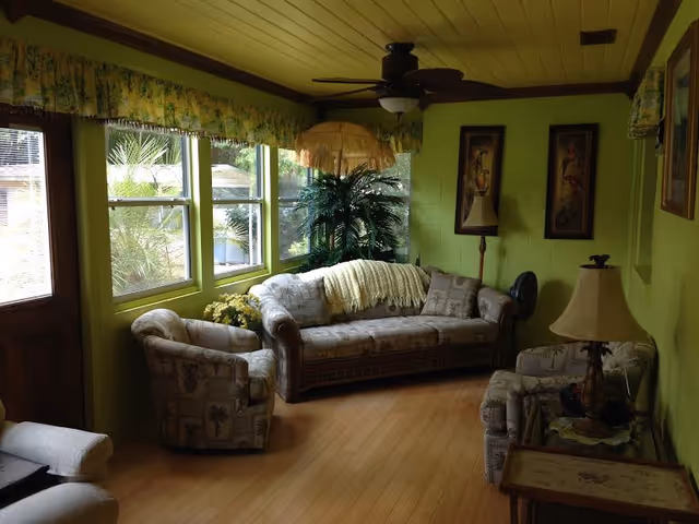 A cozy living room with light green walls and ceiling, featuring a patterned sofa and two matching armchairs. There is a ceiling fan with a light fixture, a floor lamp with a fringed shade, and a table lamp on a side table. Large windows let in natural light and show greenery outside. The room has wooden flooring and decorative framed artwork on the walls.
