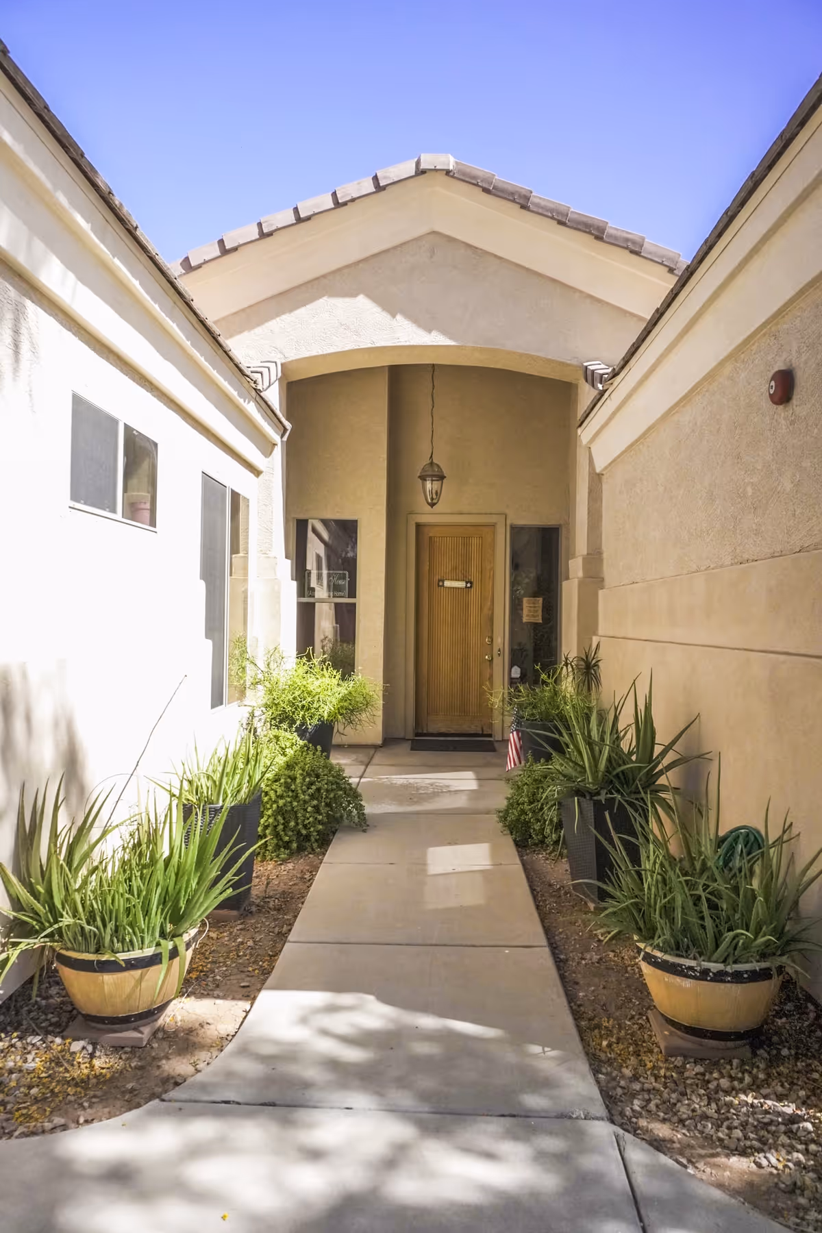 Walkway lined with potted plants leading to a covered front entrance with a wooden door and hanging light under a tiled gable roof.