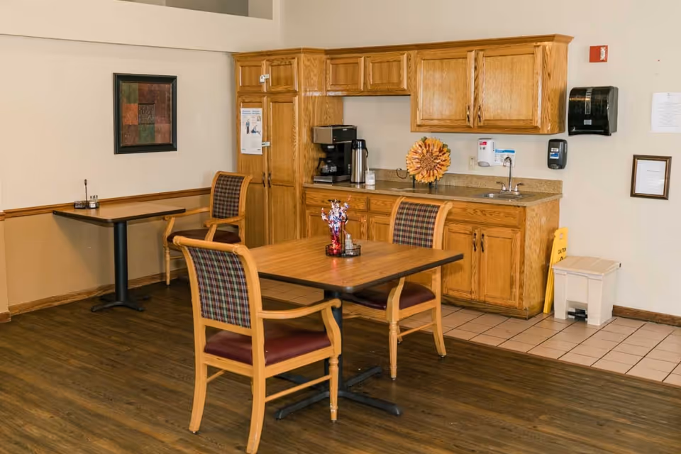Interior view of a senior living facility dining area with wooden tables and chairs featuring plaid upholstery. The room includes a kitchenette with wooden cabinets, a coffee maker, a sink, and wall-mounted dispensers. There is a framed artwork on the wall and a small decorative centerpiece on the table.
