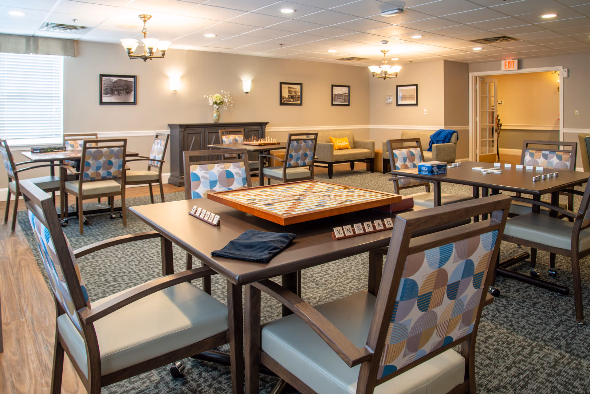 A well-lit common area in a senior living facility with multiple tables and chairs arranged for games and social activities. One table has a Scrabble board set up, and another has dominoes. The room features patterned chairs, a carpeted floor, framed pictures on the walls, a sideboard with a vase of flowers, and a seating area with sofas and cushions in the background.