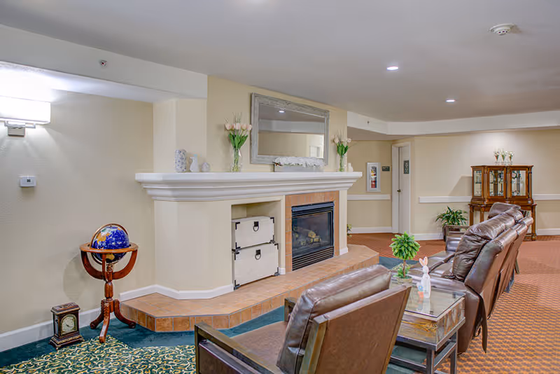 A cozy senior living facility common area with a fireplace, two brown leather chairs, a glass coffee table with decorative items, a globe on a wooden stand, and a wooden cabinet in the background. The room has beige walls, carpeted floors, and soft lighting.
