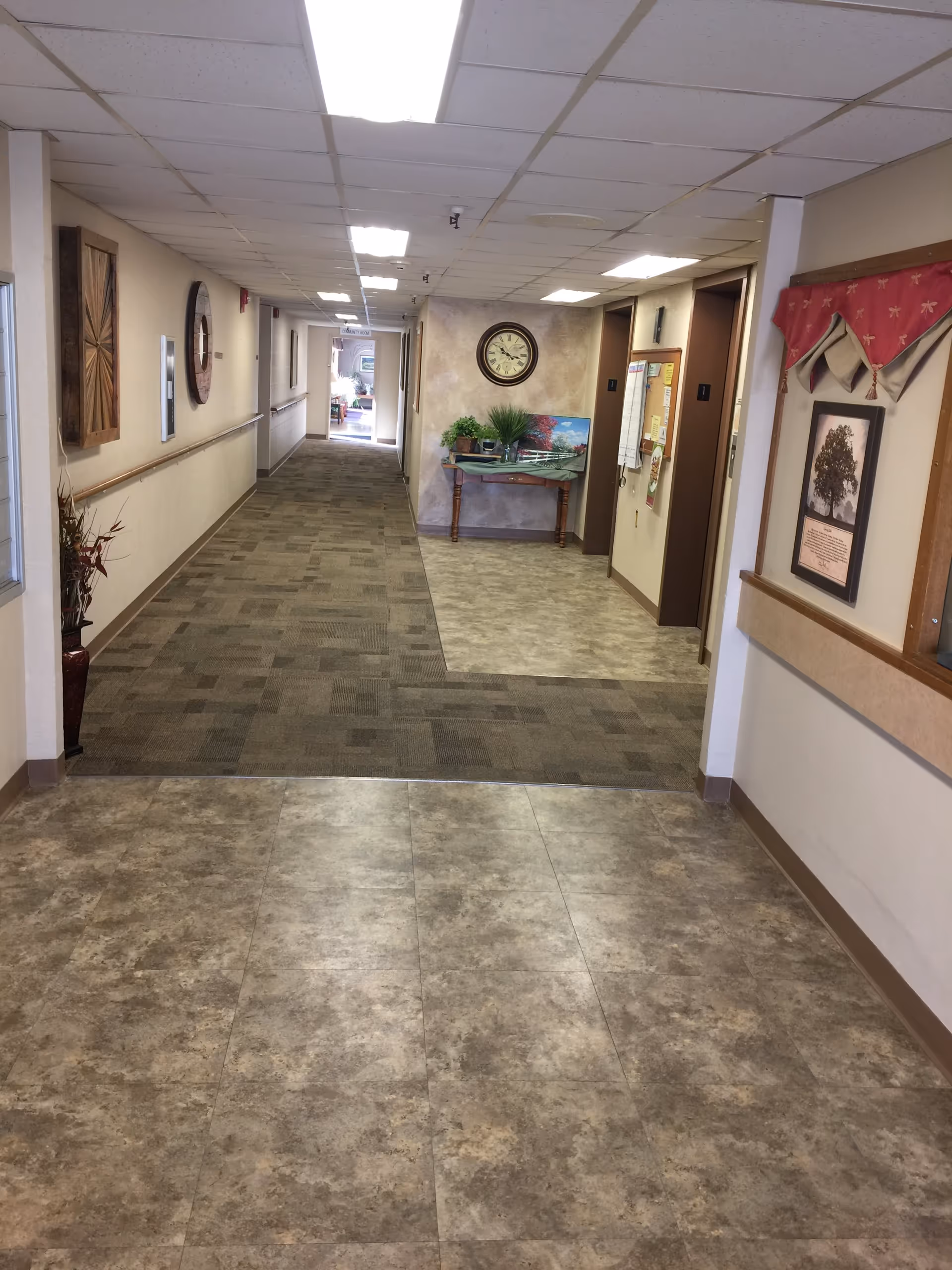 A well-lit interior hallway in a senior living facility with tile foreground, a carpeted corridor, wall art, and a table with plants and a clock at the far end.