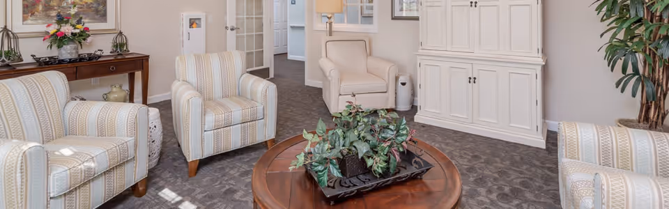 A cozy senior living room with four upholstered armchairs arranged around a round wooden coffee table with a leafy green plant centerpiece. The room features a patterned carpet, a white cabinet against the wall, a wooden console table with decorative items and flowers, and a large potted plant in the corner.
