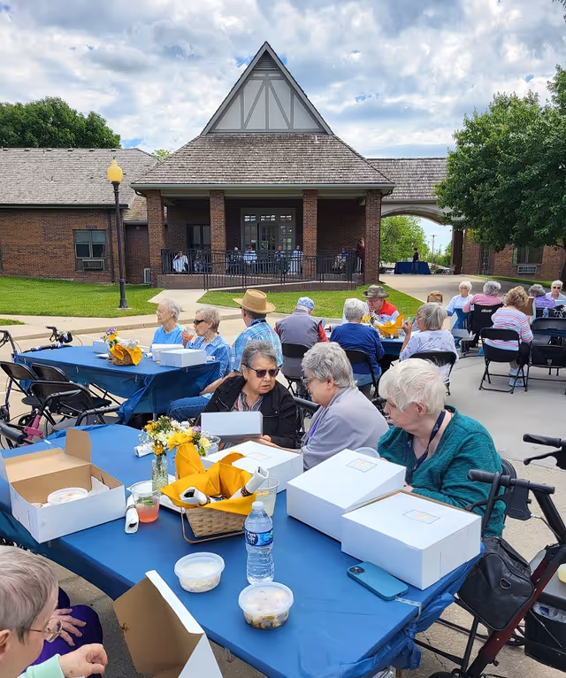 Seniors gathered at outdoor tables with boxed lunches in front of a brick community building under a partly cloudy sky.